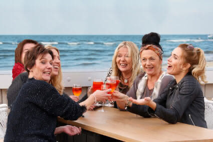 Sechs Frauen sitzen auf einer Terasse mit Meerblick in Egmond aan Zee und trinken gemeinsam Aperol Spritz