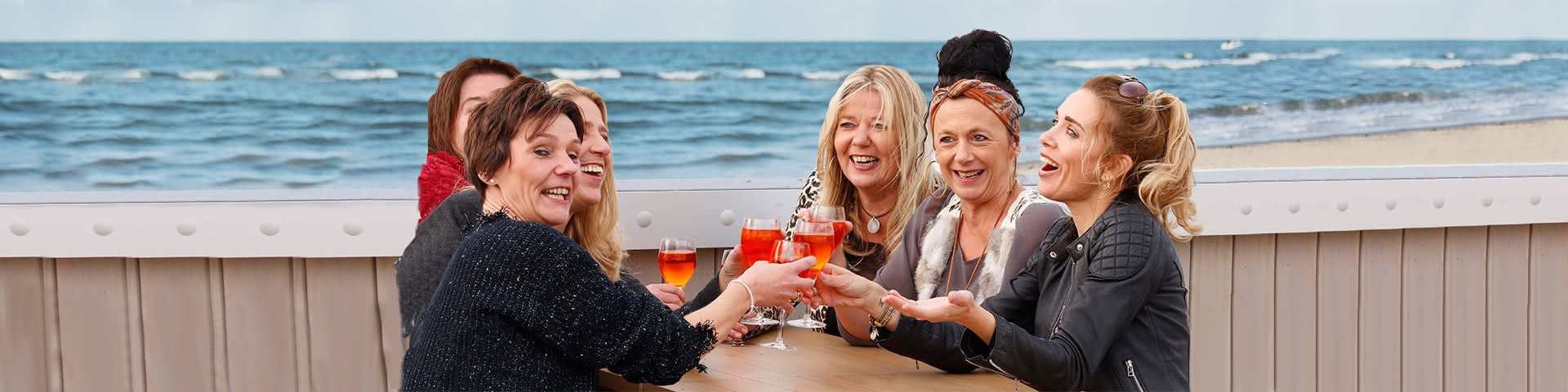 Sechs Frauen sitzen auf einer Terasse mit Meerblick in Egmond aan Zee und trinken gemeinsam Aperol Spritz