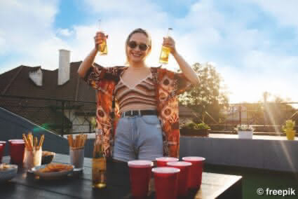 eine Frau steht auf einer Dachterrasse und hält zwei Flaschen Salitos Bier in die Höhe. Auf dem Tisch stehen Becher und Snacks